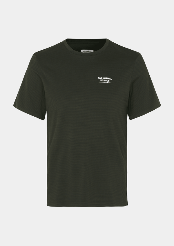 Front view of the Pas Normal Studios Men’s Balance Short Sleeve Top in the color dark olive green. Knit construction and split hem vents. Pas Normal Studios text logo in white on front left breast of shirt. 